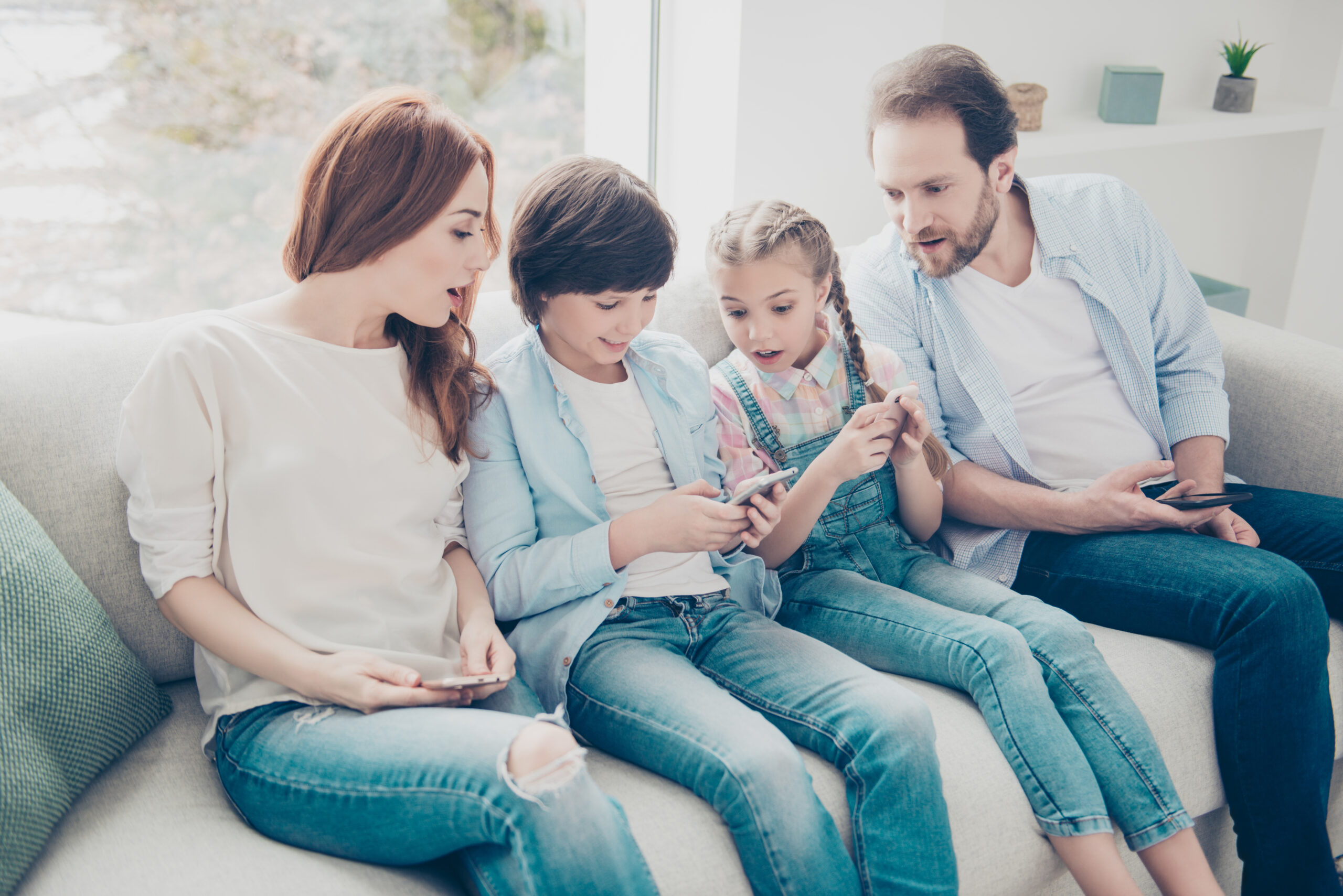 Portrait of attractive family sitting on sofa in casual outfit, mom sister and dad looking in son's smart phone with astonished impressed reaction. Social networks online connection internet concept