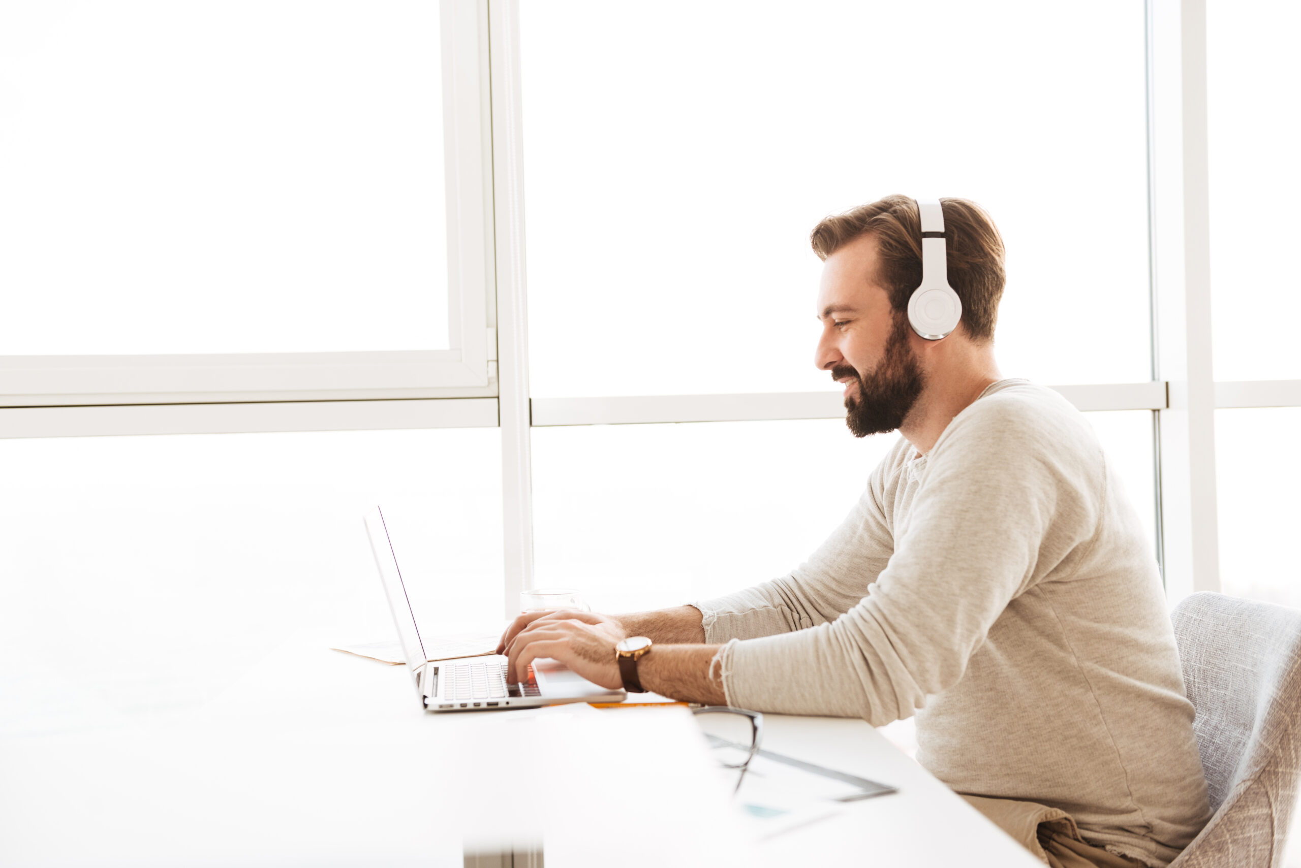 Image in profile of modern guy 30s using white headphones and communicating on laptop while resting in hotel apartment