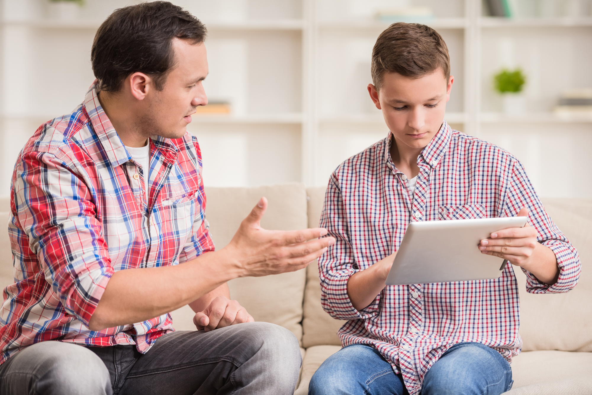 Boy sitting at sofa and using digital tablet while his father talking to him.